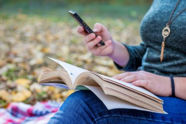 Doğadaki kız elinde bir kitap ve cep telefonu tutuyor.