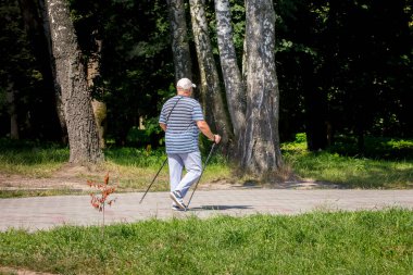 Yaşlı ve engelli bir adam sopa yardımıyla parkta dolaşıyor.
