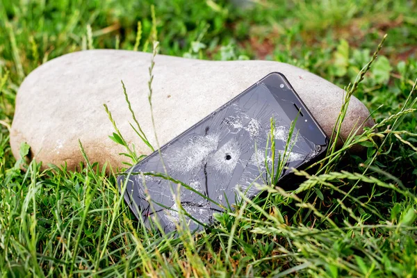 A phone with a broken touch screen and a hole from a bullet in a grass near a large stone