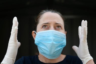 Çaresiz orta yaşlı bir kadın (30-40 yaşlarında) maske takıyor ve ellerini havaya kaldırıyor.