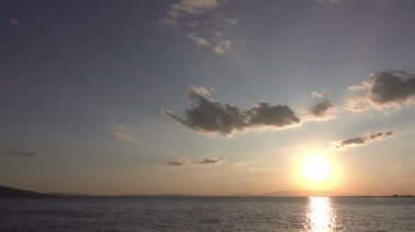 Yumuşak bulutların yavaşça solan güneşe doğru hareket ettiğini gösteriyor. Güneş 'in altın-turuncu ışınları okyanusa yansıyor.