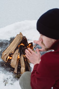 Kamp ateşi ellerine ısınma erkek bir kış döneminde kar karşı çevrili yakınındaki donmuş gölün. Kavramı macera aktif tatiller spor hiking açık 