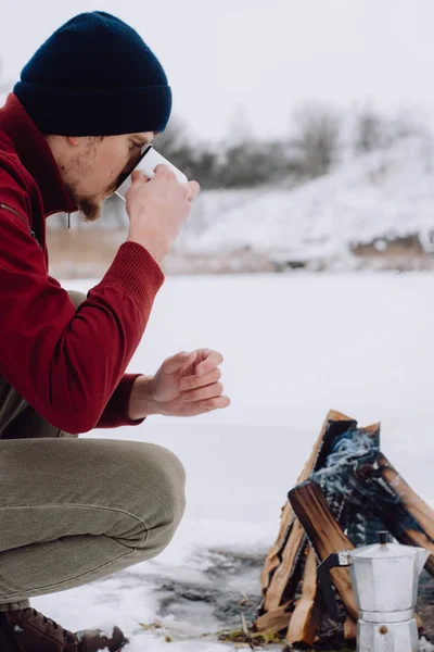 Traveling man drinks coffee by the camp fire an in winter time, surrounded by snow against the near of the frozen lake. Concept adventure active vacations outdoor hiking sport
