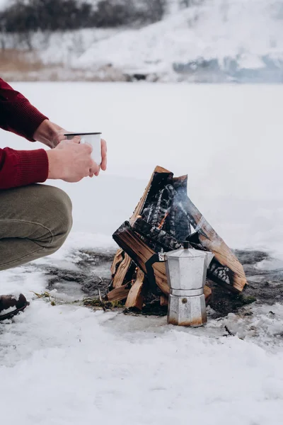 Traveling man with coffee sitting by the camp fire an in winter time, surrounded by snow against the near of the frozen lake. Concept adventure active vacations outdoor hiking sport