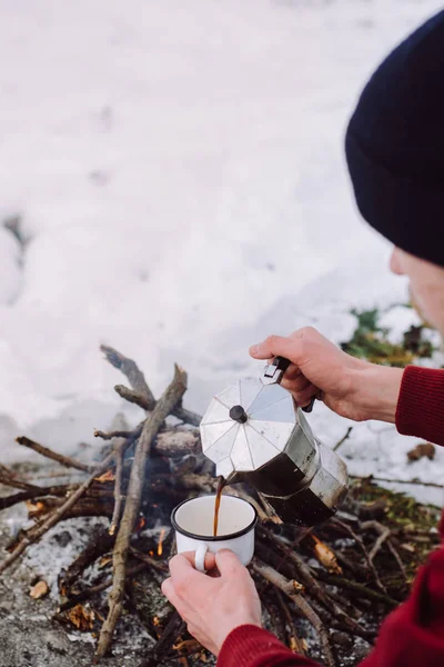 Traveling man a sits near camp fire an in winter time and pours itself hot coffee . Concept adventure active vacations outdoor. Winter camping