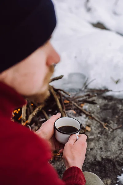 tourist is sitting near camp fire with a cup of hot coffee an in winter time. Concept adventure active vacations outdoor. Winter camping