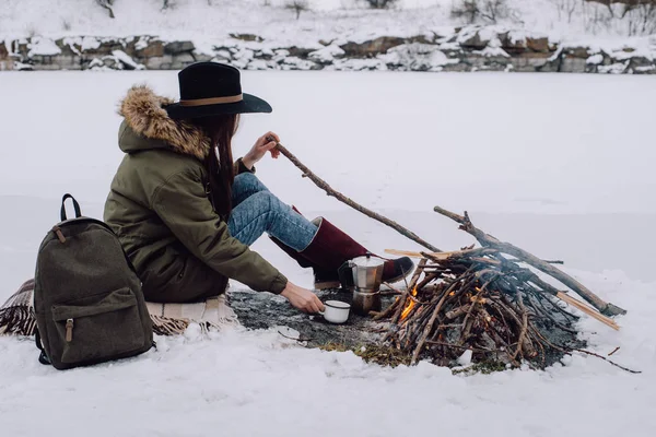 Girl tourist in a hat sits near at campfire an in winter time, surrounded by snow against the near of the frozen lake. Concept adventure active vacations outdoor. Winter camping
