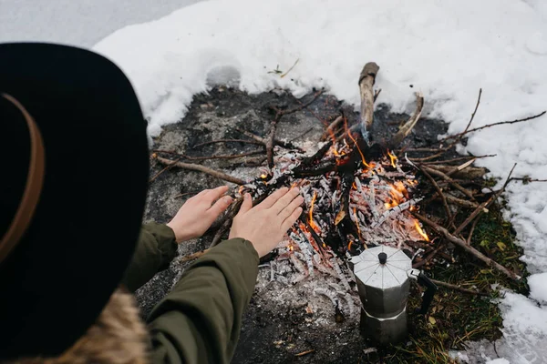 Girl in the hat warms hands near a fire in winter. Concept adventure active vacations outdoor. Winter camping