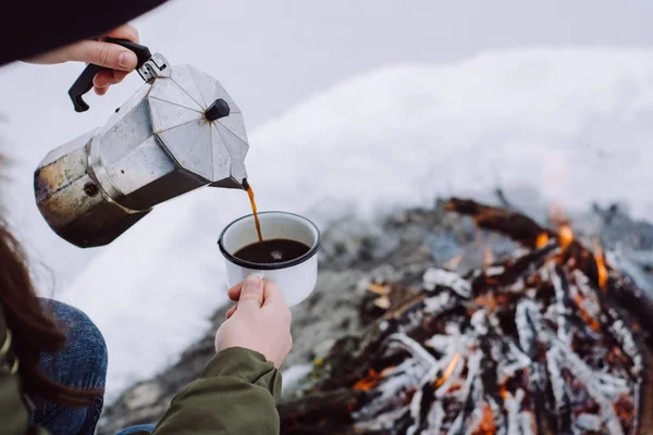 Traveling-woman  a  sits near camp fire an in winter time and pours itself hot coffee . Concept adventure active vacations outdoor. Winter camping