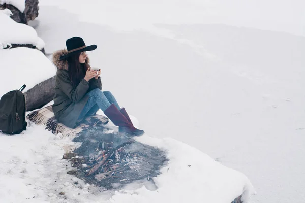 Girl in a hat sits near camp fire with a cup of hot coffee an in winter time. Concept adventure active vacations outdoor. Winter camping