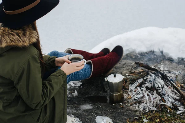 Traveling girl sits near camp fire with a cup of hot coffee an in winter time. Concept adventure active vacations outdoor. Winter camping