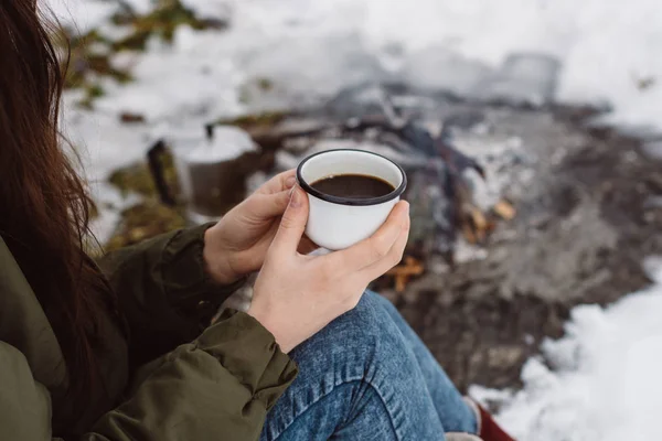 Traveling girl sits near camp fire with a cup of hot coffee an in winter time. Concept adventure active vacations outdoor. Winter camping