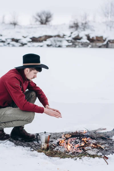 Winter camp. Tourist in hat cook coffee  and warms hands near a fire  an in winter time near of  frozen lake. Concept adventure active vacations outdoor.