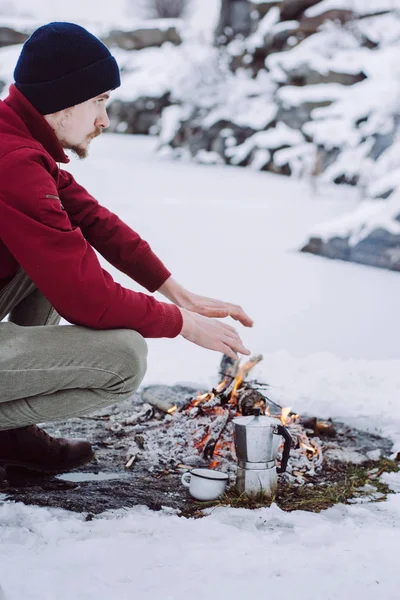 Winter camp. Man warms hands near a fire and cook coffee  the near of the frozen lake. Concept adventure active vacations outdoor.