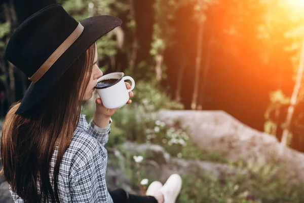 Beautiful girl in the hat drinks coffee on top of the mountain and enjoying the sunrise . Concept adventure active vacations outdoor in the mountains. Summer camp