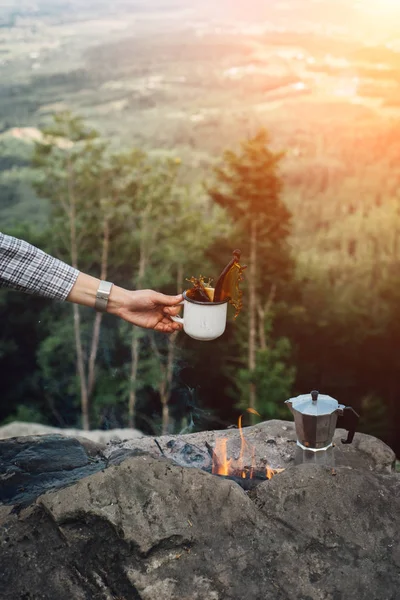 Girls hand holds a hot cup of coffee near the campfire on  background of nature during the sunset . Concept adventure active vacations outdoor. Summer camp.