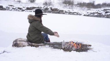 Şapkalı kız ellerini yangın yanında kışın donmuş gölün arka plan üzerinde ısıtıyor. Kavramı macera aktif tatiller açık. Kış kamp