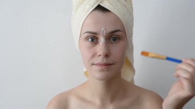Yüzünü bir fırça sonra bir duş ile krem uygulayarak banyo havlusu ile sarılmış çekici genç kadın. Cilt bakım ve güzellik kavramı. Kadın yüzüne evde yüz maskesi uygulama.
