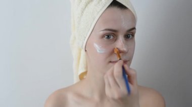Yüzünü bir fırça sonra bir duş ile krem uygulayarak banyo havlusu ile sarılmış çekici genç kadın. Cilt bakım ve güzellik kavramı. Kadın yüzüne evde yüz maskesi uygulama.