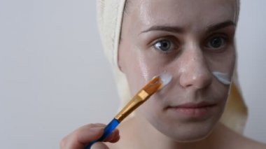 Yüzünü bir fırça sonra bir duş ile krem uygulayarak banyo havlusu ile sarılmış çekici genç kadın. Cilt bakım ve güzellik kavramı. Kadın yüzüne evde yüz maskesi uygulama.