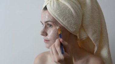 Yüzünü bir fırça sonra bir duş ile krem uygulayarak banyo havlusu ile sarılmış çekici genç kadın. Cilt bakım ve güzellik kavramı. Kadın yüzüne evde yüz maskesi uygulama.
