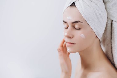 Güzel genç kadın banyo havlusu ile sarılmış krem yüzüne alır. Cilt bakım kavramı. Mükemmel sağlık temiz cilt ile çarpıcı beyaz kadın. 