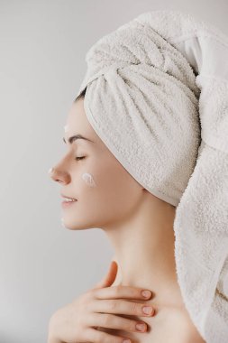 Güzel genç kadın banyo havlusu ile sarılmış krem yüzüne alır. Cilt bakım kavramı. Mükemmel sağlık temiz cilt ile çarpıcı beyaz kadın. 