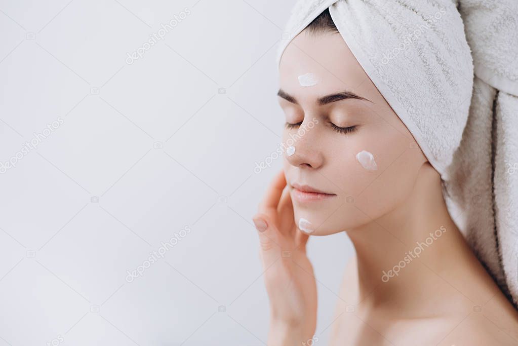 Beautiful young woman wrapped with bath towel gets cream in the face. Skin care concept. Stunning caucasian woman with perfect health clean skin. 