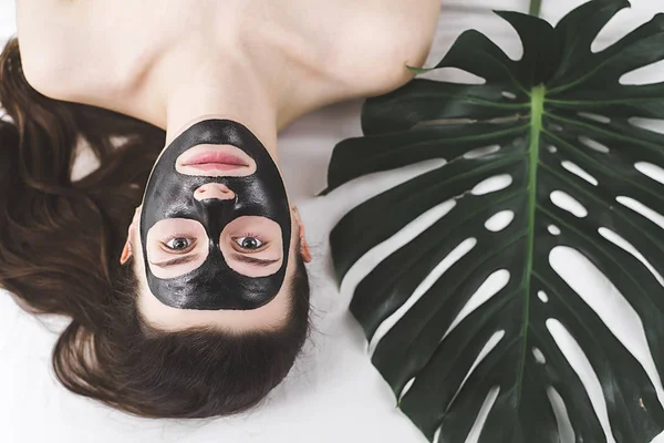 Çekici kızı aç gözlerini ve yatakta yatan yaprak yeşil siyah bir maske yüzüne karşı uygulanır. Kadın siyah bir yüz maskesi ile rahatlatıcı. Kavram sağlık ve güzellik . 