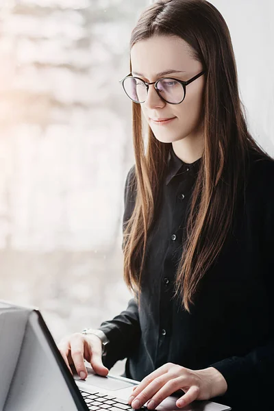 Girl in the glasses is working on a portable computer at home. Attractive young woman sits near window and typing messages on a laptop.