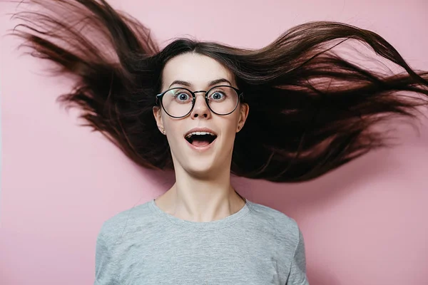 Giovane ragazza in occhiali con i capelli volanti in panico guarda con espressione sorpresa, si sente nervoso in situazione di stress, indossa maglietta grigia, apre la bocca con stupore, isolato sulla parete rosa — Foto Stock