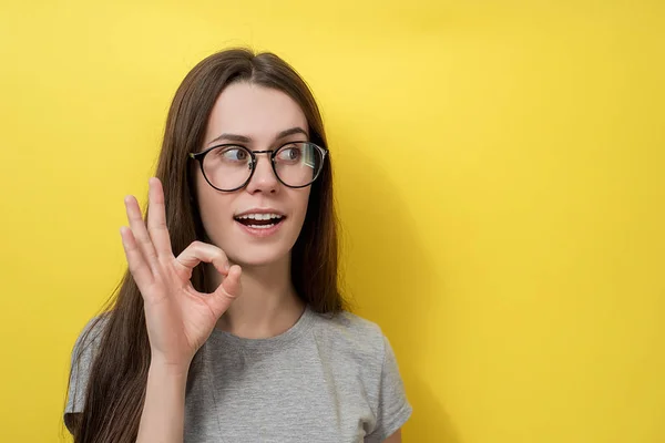 Ragazza ottimista in occhiali mostra un gesto eccellente, con sguardo stupito, vestito in grigio T-shirt, isolato su sfondo giallo. Femmina dimostra segno ok — Foto Stock