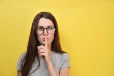 Şaşkın ifade, gözlüklü genç kadın hareketi kapalı sessiz yapar, ön plana tutan dudaklar, birisi onun sırrını açığa endişeler parmağında duruyor sarı arka planı