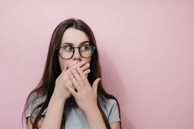 Hayrete çılgın kız bakışları korkmuş ifade, iki eliyle kapakları ağız ile gri T-shirt, pembe bir arka plan karşı pozlar giyer. İnsanlar ve duygu kavramı