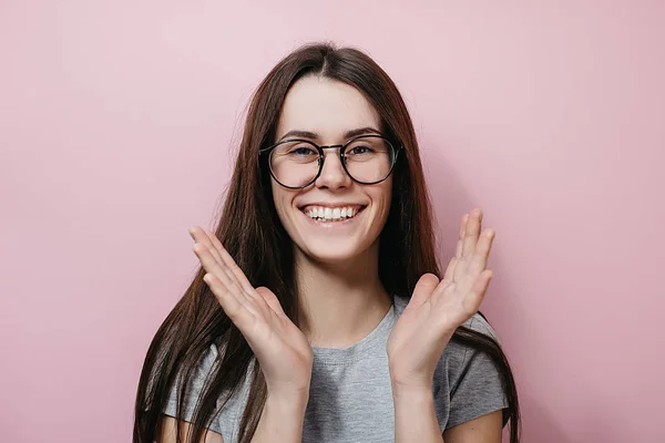 Piacevole e gioiosa giovane donna europea sorride positivamente, tiene le mani vicino alle guance, indossa occhiali, vestita con maglietta grigia, isolata su sfondo rosa. Emozioni e sentimenti concetto — Foto Stock