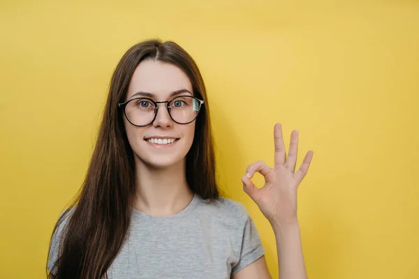 Позитивная европейская женщина в очках делает нормальный жест, носит серую футболку, наслаждается жизнью, изолирована на желтом фоне, говорит нормально, подтверждает информацию. Его отличный результат — стоковое фото