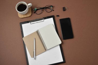 Mockup çalışma alanı boş klibi kurulu, büro malzemeleri, kalem, not defteri, smartphone, kahve fincanı ve kahverengi zemin üzerine gözlük ile. Düz yatıyordu, en iyi görünüm, şık konsept