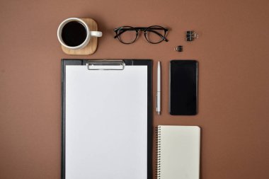 Mockup çalışma alanı boş klibi kurulu, büro malzemeleri, kalem, not defteri, smartphone, kahve fincanı ve kahverengi zemin üzerine gözlük ile. Düz yatıyordu, en iyi görünüm, şık konsept
