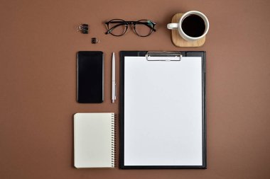Mockup çalışma alanı boş klibi kurulu, büro malzemeleri, kalem, not defteri, smartphone, kahve fincanı ve kahverengi zemin üzerine gözlük ile. Düz yatıyordu, en iyi görünüm, şık konsept