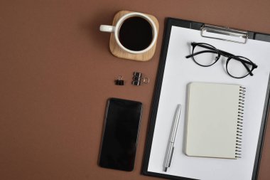 Mockup çalışma alanı boş klibi kurulu, büro malzemeleri, kalem, smartphone, not defteri, kahve fincanı ve kahverengi zemin üzerine gözlük ile. Düz yatıyordu, en iyi görünüm, şık konsept