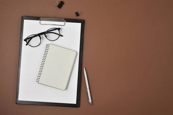 Preencha o espaço de trabalho com placa de clipe em branco, material de escritório, caneta, bloco de notas e óculos no fundo marrom. Flat lay, vista superior, espaço de cópia — Fotografia de Stock