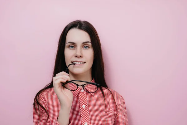 Ritratto di felice giovane donna tiene gli occhiali vicino alle labbra, si rallegra di ricevere complimenti, vestito con abiti casual, isolato su sfondo rosa con spazio copia per la tua pubblicità — Foto Stock