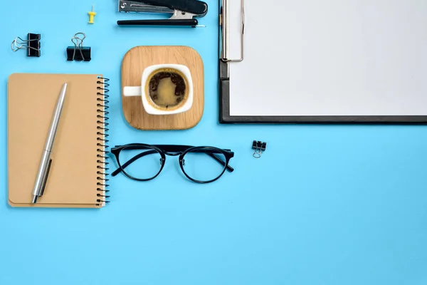 Office workspace with blank clip board, office supplies, pen, notepad, eyeglasses, stapler, coffee cup and spectacles on blue background. Flat lay, top view, stylish concept