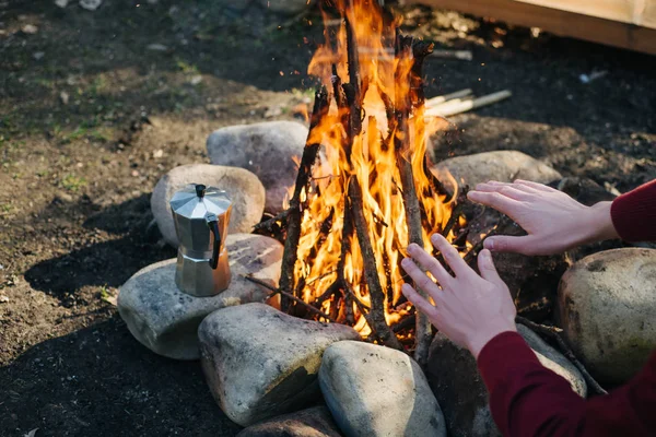 Outdoor horizontal image of traveler warming his hands by the campfire an in summer time. Rear view of traveler man in red jacket, sitting near to bonfire, after hiking. Travel concept