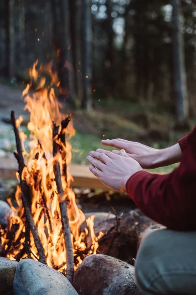 Περικομμένη εξωτερική εικόνα του νεαρού άντρα που ζεσταίνει τα χέρια του από την κοντινή φωτιά. Πίσω όψη του ταξιδιώτη με κόκκινο μπουφάν, κάθεται κοντά στη φωτιά, μετά την πεζοπορία. Σχέδιο ταξιδίου — Φωτογραφία Αρχείου