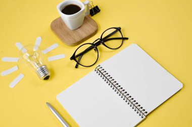 Üst görünüm düz bir sarı renkli kağıt arka plan minimal tarzı üzerinde kalem, Not defteri, Gözlük, fincan kahve ve ampul ile çalışma masası tarzı tasarım ofis malzemeleri yatıyordu. Konsept beyin fırtınası