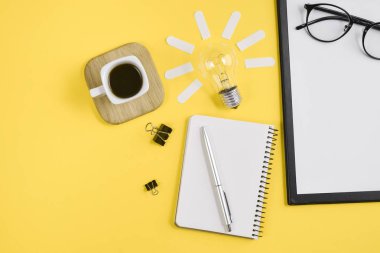 Üst görünüm düz bir sarı renkli kağıt arka plan minimal tarzı üzerinde kalem, Not defteri, Gözlük, fincan kahve ve ampul ile çalışma masası tarzı tasarım ofis malzemeleri yatıyordu. Konsept beyin fırtınası