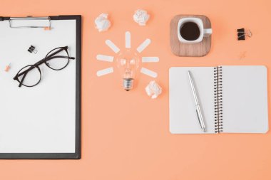 Düz boş klip Board, ofis malzemeleri, kalem, Not defteri, Gözlük, kahve fincan, turuncu arka planda ampul ile dağınık masa üst görüntü beyin fırtınası mali planlama yatıyordu.