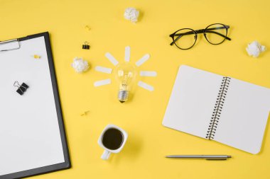 Düz boş klip Board, ofis malzemeleri, kalem, Not defteri, Gözlük, kahve fincan, sarı arka planda ampul ile dağınık masa üst görüntü beyin fırtınası mali planlama yatıyordu.