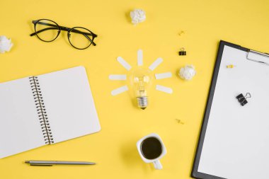 Boş klip panosu, ofis malzemeleri, kalem, Not defteri, Gözlük, kahve bardağı ve sarı arka planda ampul ile Office çalışma alanı. Konsept beyin fırtınası ve yeni fikir